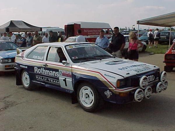 A classic Audi rally car with a Rothmans livery, featuring multiple fog lights, parked at a car event with a crowd of people in the background.