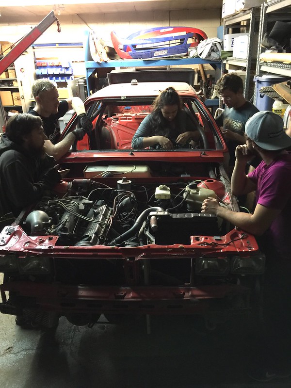29073839983_bb1d7a8294_c Five people working on and around a red car chassis in a dimly lit garage with tools and automotive parts