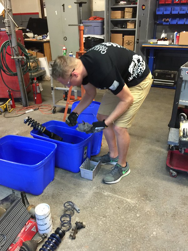 29698687695_834d51d71f_c Person wearing black shirt and khaki shorts handling car parts near blue bins in a workshop