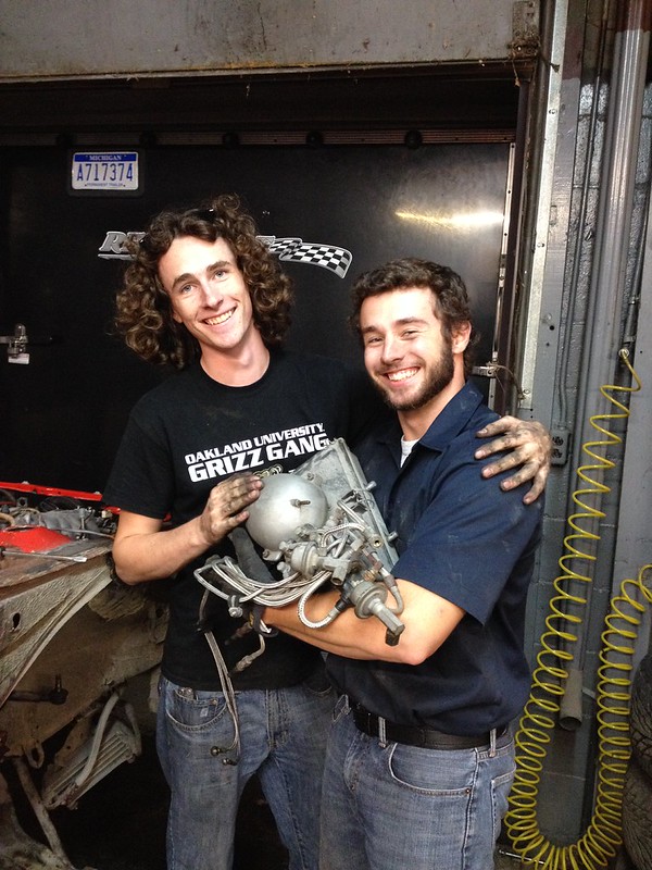 29824994503_b5a9746b44_c Two young men smiling and holding a mechanical car part in a garage with tools and equipment around
