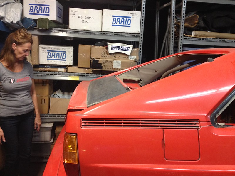29846206625_980aef7e9d_c A woman standing next to a red vintage car in a storage area with shelves filled with boxes
