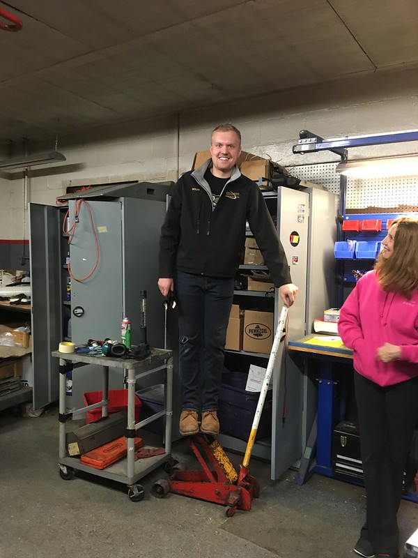 30118942214_9596583b2a_c A man standing on a red hydraulic floor jack in a workshop while a woman in a pink hoodie watches nearby