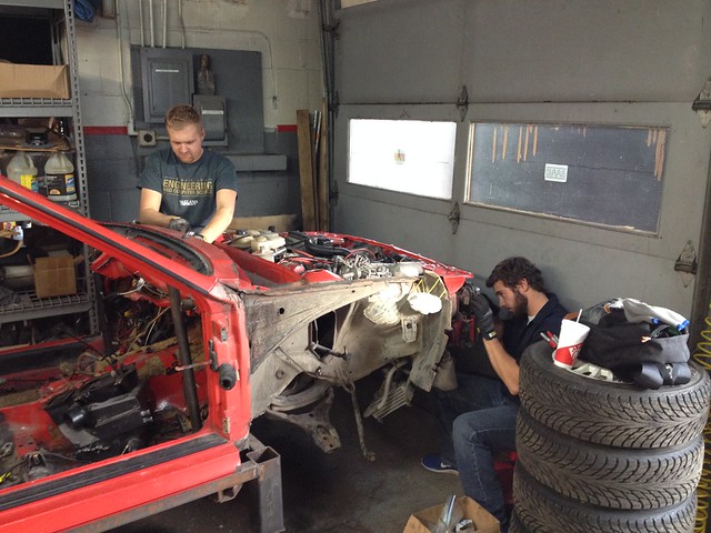 30420932776_9f41cca2e0_z Two men working on disassembled red car frame inside a garage with stacked tires nearby