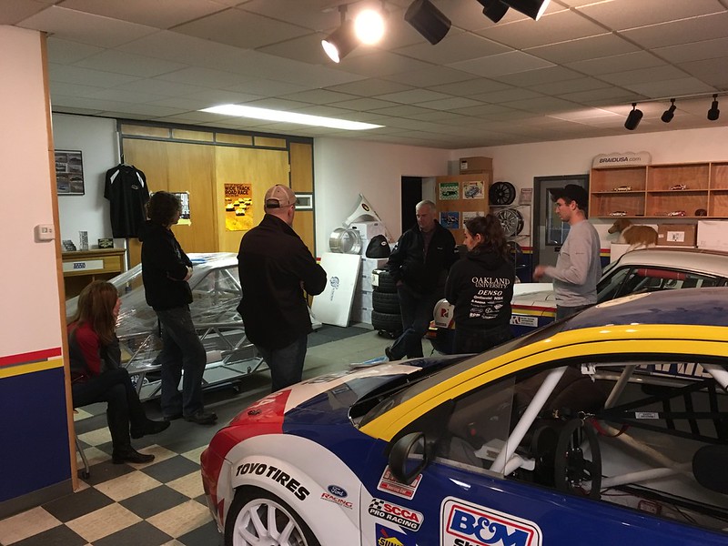 30630720135_a8a0bd4b93_c Group of people standing in a garage discussing near race cars and automotive equipment