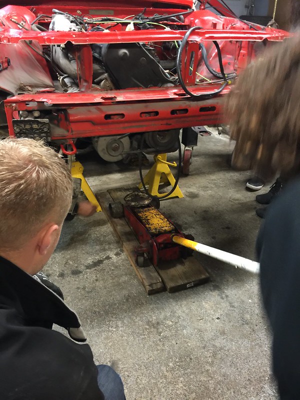 30693276601_46b1e5a658_c People working on a red car lifted by jack stands in a garage with a floor jack beneath it