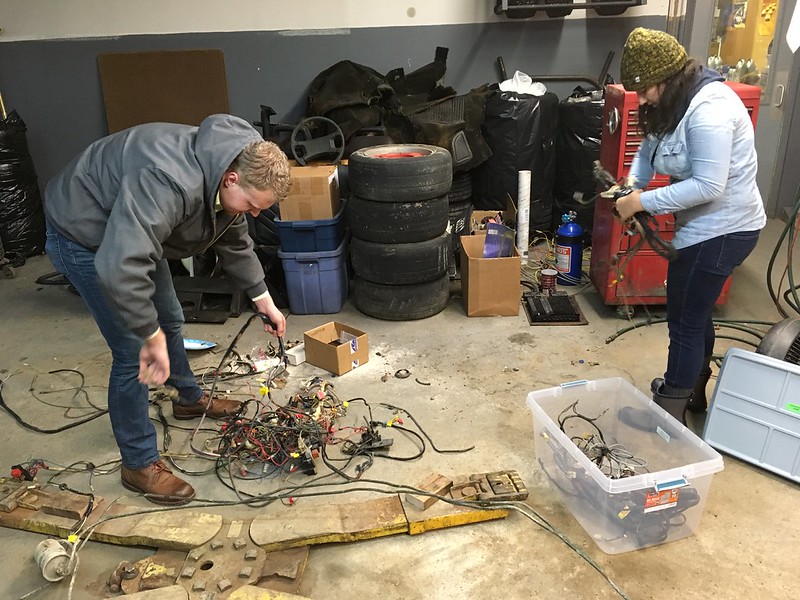 Two people sorting a pile of wires and electronic components in a workshop with tires and storage bins in the background
