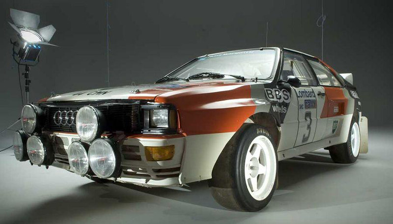 A vintage Audi rally car with multiple headlights, featuring a two-tone paint scheme and race number on the side, positioned in a studio setting.