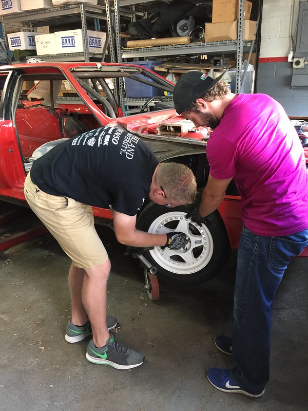 img Two men working together to install or adjust a tire on a red car inside a garage