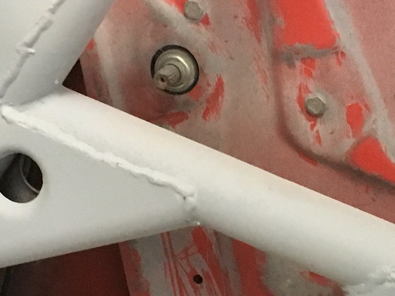 Close-up of a metal frame with visible welds and a bolt near a round opening, featuring a partially obscured electrical or mechanical component.