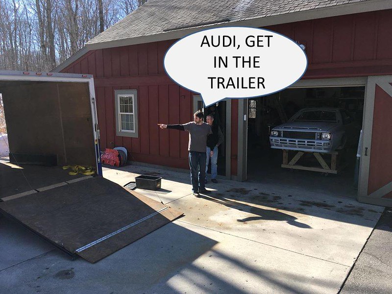 Two men standing outside a red barn, one pointing towards a trailer while the other looks on. A speech bubble above the first man reads, 'AUDI, GET IN THE TRAILER.' A car is visible inside the barn.