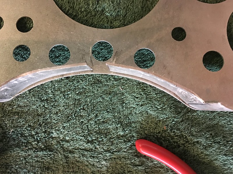 Close-up view of a metal piece with circular cutouts and a curved edge, lying on a green textured surface, next to a red tool.