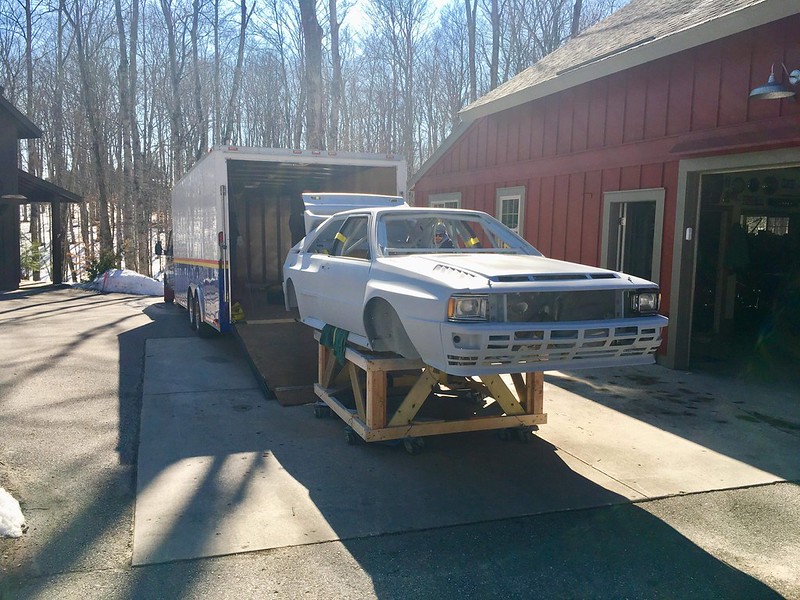 A partially assembled white racing UR Quattro on a wooden platform in front of a trailer and a red building, surrounded by trees.