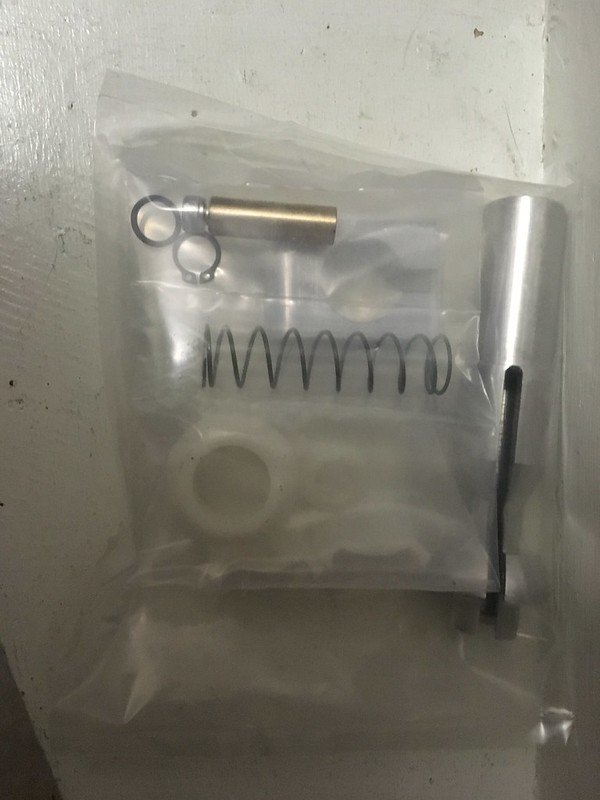 A clear plastic bag containing automotive or mechanical components including a spring, a cylindrical metal part, a pin, and plastic washers.