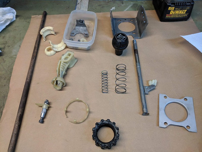 Assorted mechanical parts laid out on a work surface, including rods, springs, and plastic components, along with a container holding a piece.