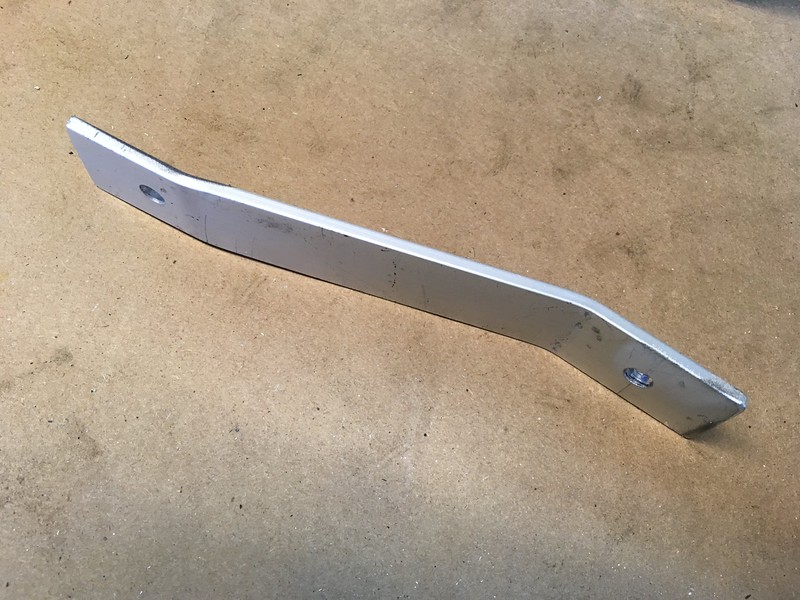 A metallic bracket with a slight bend, featuring two holes for mounting, resting on a brown surface.