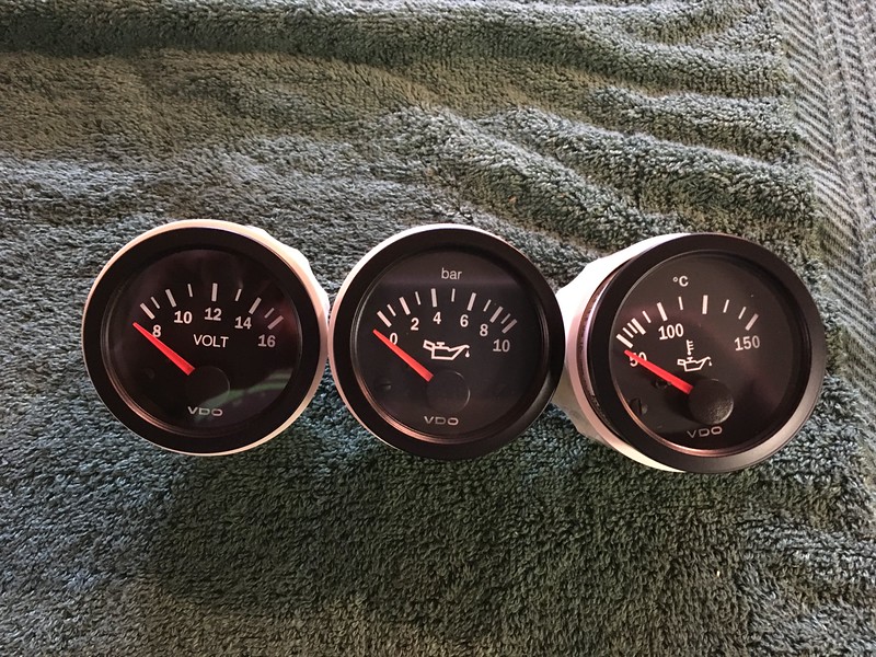 Three automotive gauges displaying voltage, oil pressure (in bar), and oil temperature (in Celsius) on a textured surface.