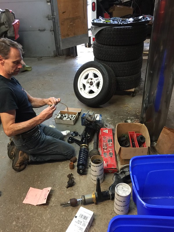 A mechanic working on UR Quattro rally parts in a workshop, surrounded by tools, tires, and disassembled components.