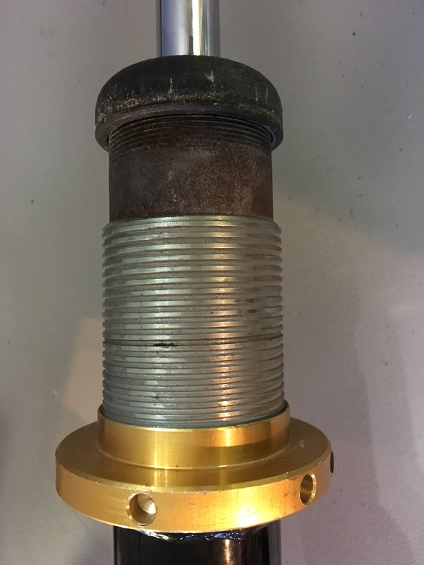 Close-up of a metal suspension coilover with a threaded body, showcasing a gold-colored base and a dark top.
