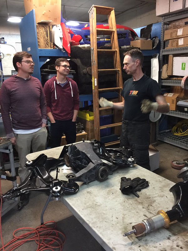 33345353653_afa26f155a_c Three men in a workshop with automotive parts on a table and shelves with boxes and tools around