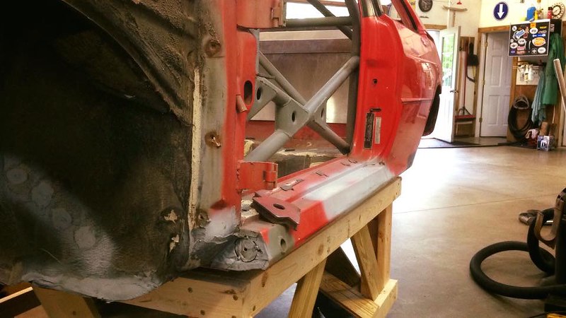 Close-up view of the side of a red car on a wooden stand, showing the exposed interior and structural components, with a garage background.