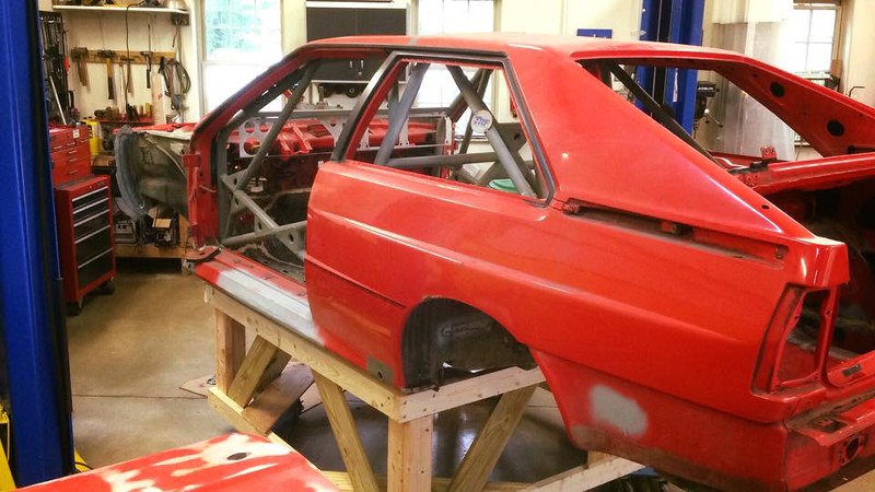 A partially disassembled red car body on a wooden frame in an automotive workshop, with tools and equipment in the background.