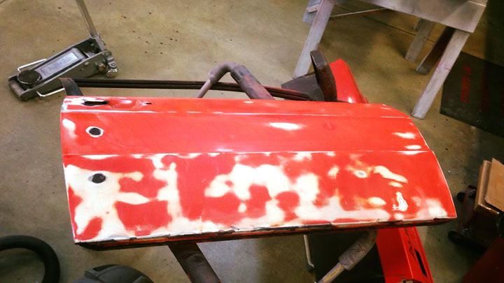 A red car door panel with some areas of primer showing through, placed on a workbench in a garage.