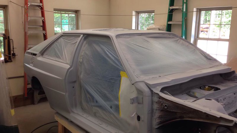 A car partially covered in plastic sheeting, positioned in a workshop, with a ladder and tools visible in the background.