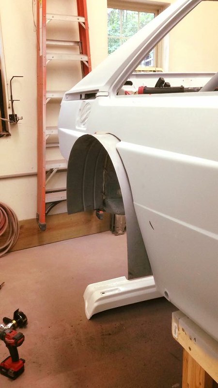 A partially assembled UR Quattro in a workshop, featuring a white car body with visible fender work and tools scattered on the floor.