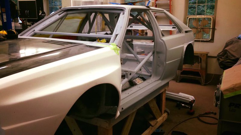 A partially assembled UR Quattro body on a wooden stand, showcasing its metallic frame and exterior paneling in a workshop environment.