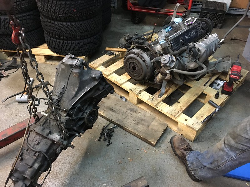 A disassembled UR Quattro engine and transmission sitting on wooden pallets in a garage, with tires in the background and various tools scattered on the floor.