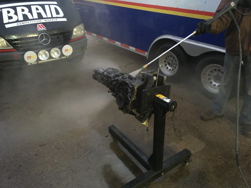 A person cleaning a UR Quattro transmission with a pressure washer, mounted on a stand, next to a vehicle and trailer.
