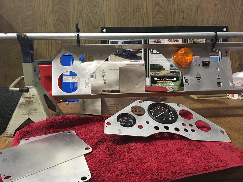An assortment of metal dashboard panels and gauges placed on a red towel, with a mounting structure in the background.