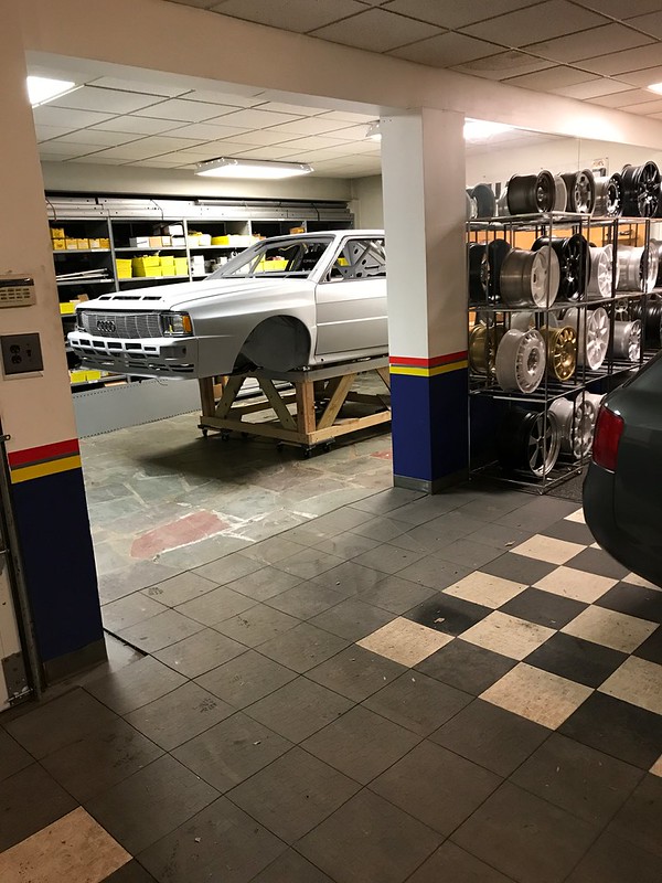 An automotive workshop featuring a partially assembled white UR Quattro on a wooden frame, surrounded by shelves of automotive parts and a display of various wheels.