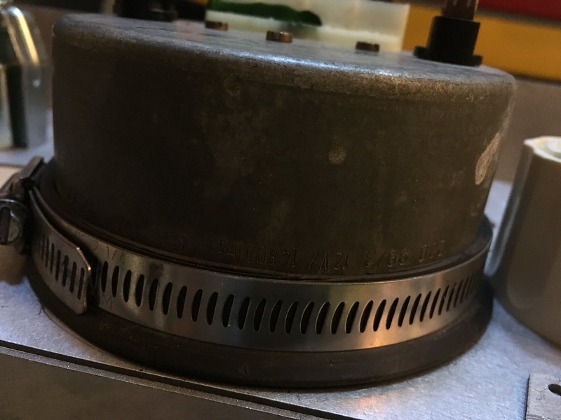 Close-up view of a metallic cylindrical object with a clamp around its base, showing wear and inscriptions, placed on a flat surface with other equipment in the background.