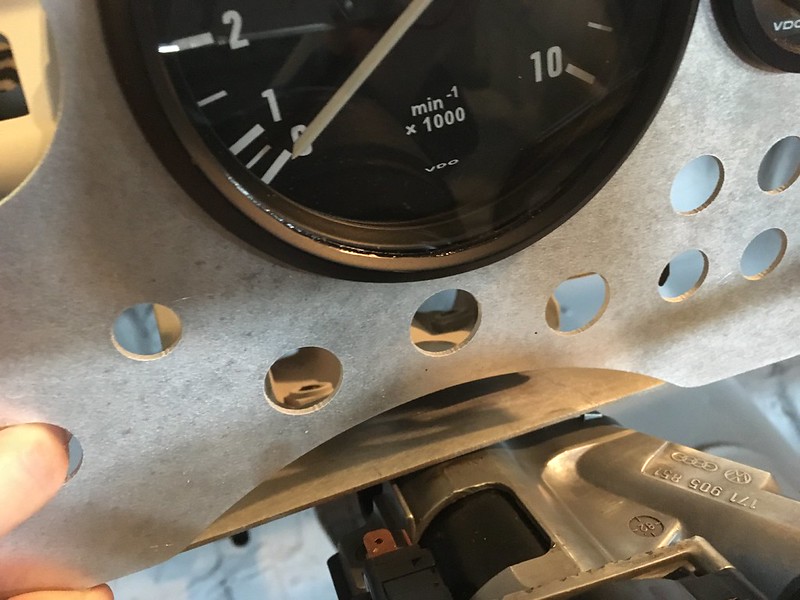 Close-up image of a UR Quattro rally dashboard gauge surrounded by a perforated metal cover, with various holes visible and a hand holding the cover.