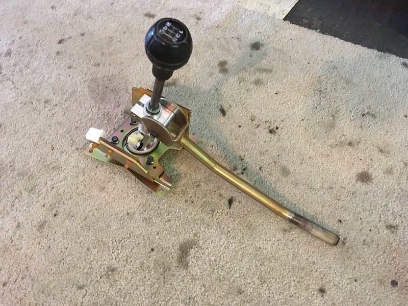 A UR Quattro rally gear shift lever assembly mounted on a base, featuring a black shift knob and a metal rod, positioned on a light-colored carpet.