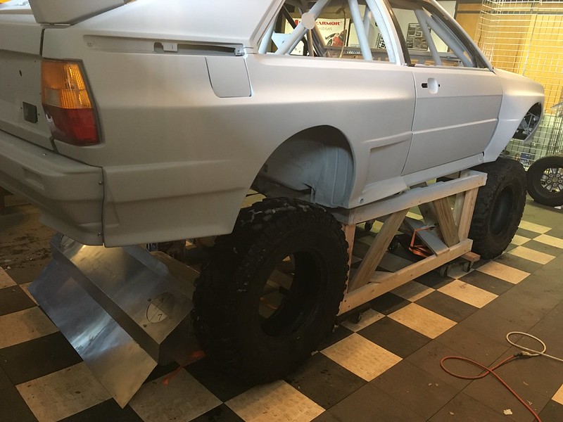 A modified UR Quattro chassis elevated on wooden stands, featuring oversized tires and a plow attachment at the front, set against a checkered floor.