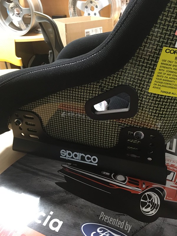Close-up view of a Sparco racing seat, showcasing its textured fabric and mounting bracket, with a warning label visible.