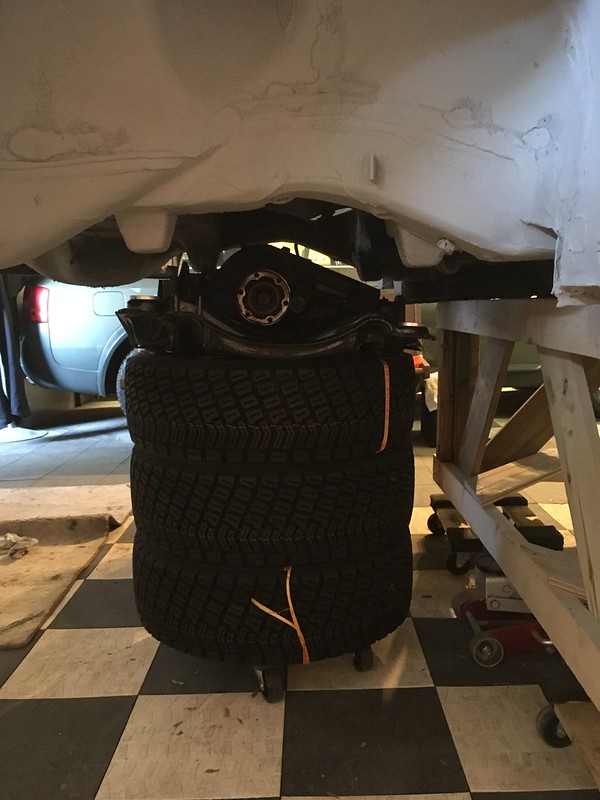 Underneath a UR Quattro , a differential assembly is supported on top of three stacked tires, all resting on a checkered floor in a garage setting.
