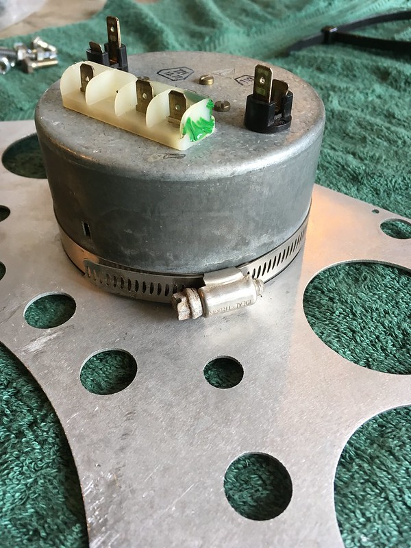A cylindrical metal component with electrical connectors on top, mounted on a circular metallic base with holes, resting on a green cloth.