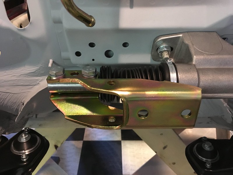 Close-up of a metallic Quattro rally steering rack mounting bracket with bolts attached, set against a white background and checkered floor.