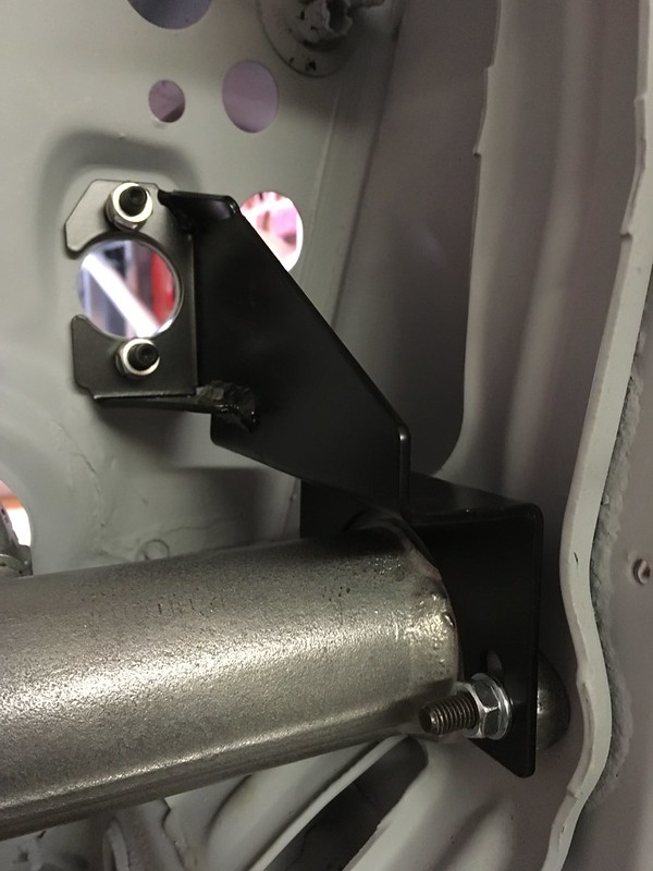 Close-up image of a metal bracket attached to a cylindrical metal pipe, viewed from an angle showing mounting holes and screws.