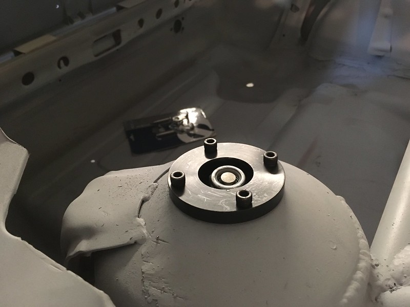 Close-up of a black metal component with six threaded holes, positioned on a gray surface, possibly part of a car's suspension system.