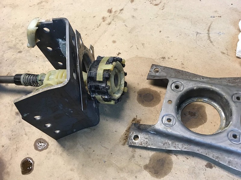 A mechanical assembly comprising a metal bracket with visible screws and a cylindrical component featuring a yellow and black design, placed on a workbench with some oil stains.
