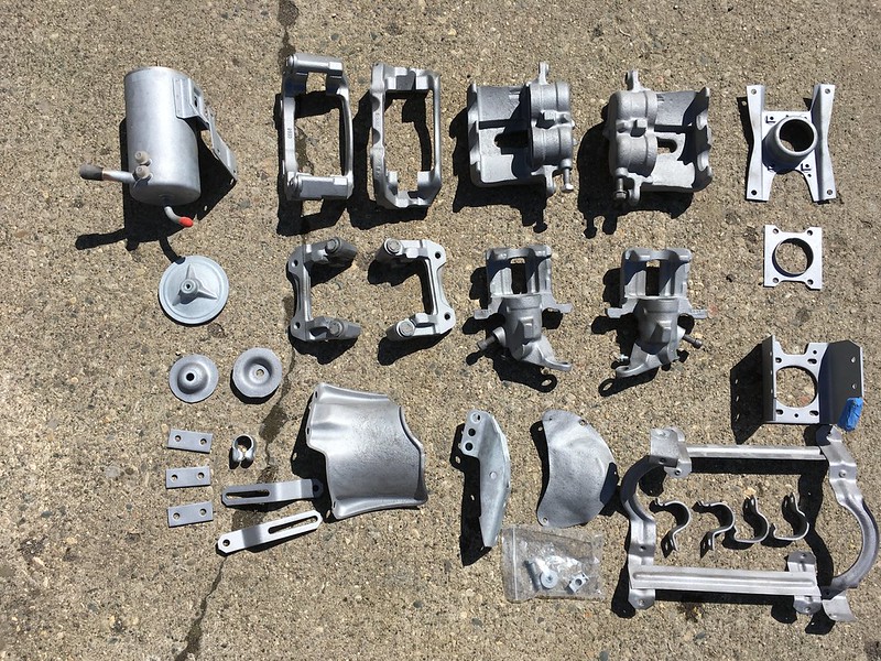 An assortment of shiny, silver automotive parts arranged on a concrete surface, including brackets, casings, and connectors.