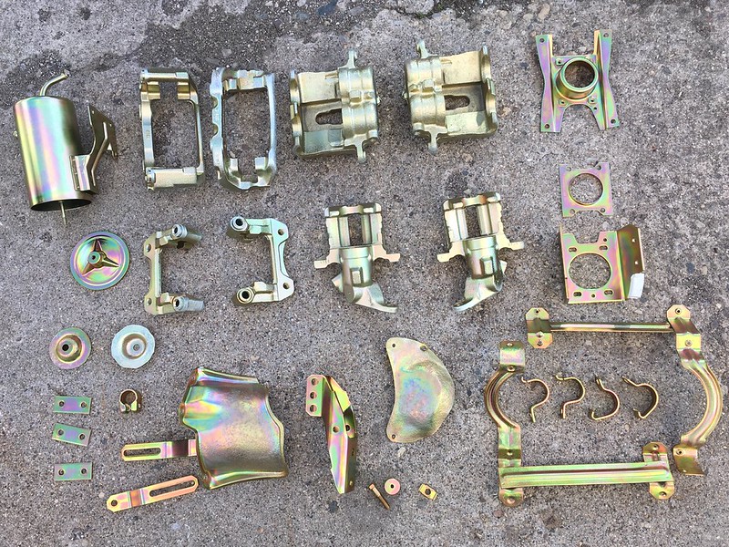 Arrangement of various metallic mechanical parts on a concrete surface, featuring a mix of shapes and sizes with a shiny, rainbow-colored finish.