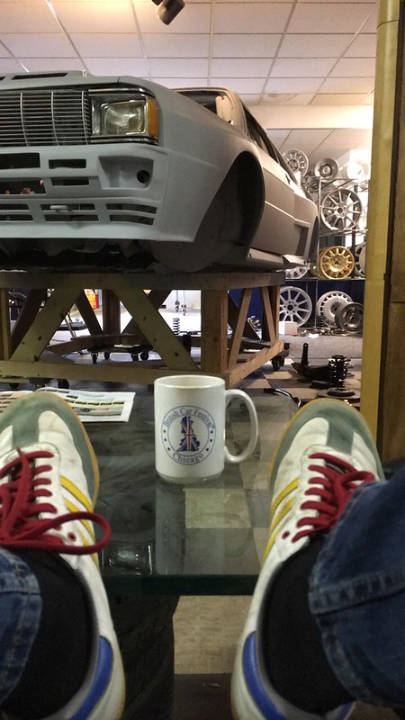 A person's feet in colorful sneakers resting on a glass table in a workshop, with a partially assembled UR Quattro rally car on a wooden platform in the background, surrounded by various car parts and wheels.