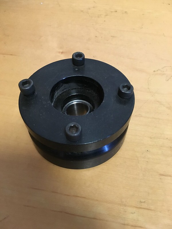 Close-up of a circular black mechanical part with a central opening and four bolts on top, placed on a wooden surface.
